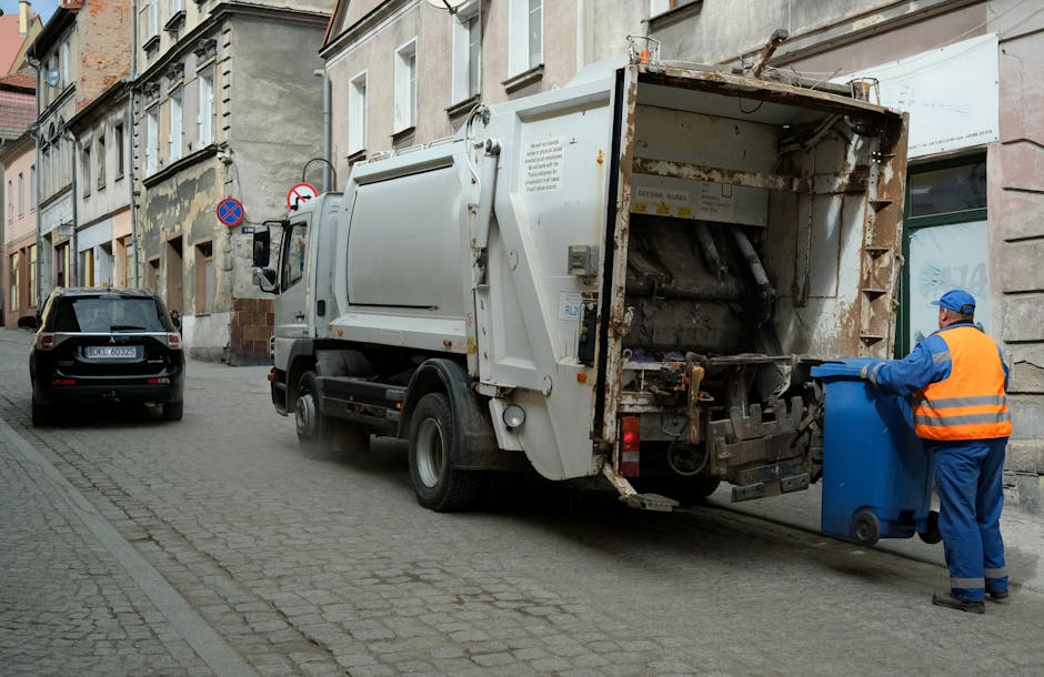 A street scene featuring a white garbage collection truck with a rusted rear panel and hydraulic lift, positioned next to a building with old, weathered facades and multiple windows. The truck is actively engaged in rubbish removal, with debris or waste material inside the open rear compartment. A worker dressed in a high-visibility orange vest and blue uniform is placing a blue wheeled bin into the truck's lifting mechanism, preparing it for empting. Parked alongside the curb is a black car, with a street sign visible behind it, indicating no stopping or parking restrictions. The scene is illuminated by natural daylight, highlighting the urban environment's textures, including cobbled pavement, aged building surfaces, and the metal surfaces of the waste vehicle. This setup reflects a standard private rubbish collection process, often utilized as an alternative to municipal waste services, aligning with the themes of independent rubbish removal and on-site clearance as discussed by Rubbish Clearance Balham.