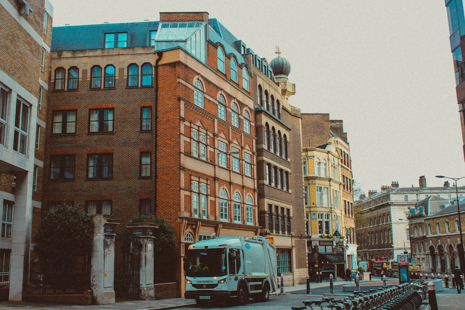 A street scene featuring a row of multi-story buildings with varied architectural styles, including brick facades, arched windows, and decorative detailing. In the foreground, a white refuse collection vehicle is parked on the pavement near a curb, partially obscuring a gated entrance with stone pillars. To the right, there are protective barriers along the sidewalk, and a street lamp is visible in the distance. The environment suggests an urban area with historic and modern buildings closely situated. Natural light illuminates the scene, highlighting the textures of the brickwork and the vehicle's smooth surface, consistent with a typical city setting involved in waste management activities. The presence of the waste collection vehicle indicates local rubbish collection or private waste disposal service activity, aligning with alternative rubbish clearance efforts by companies like Rubbish Clearance Balham, as referenced in the context of independent waste handling on busy city streets.