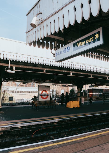The image shows a section of a train station platform beneath an elevated structure, with a large yellow 'Way Out' sign with an arrow pointing to the right suspended from the ceiling. The platform has dark paving, with several benches occupied by a few people waiting for the train. In the background, there is a white building with multiple windows, and a sign displaying the 'Golders Green' station logo with a red circle and black text. A yellow tactile paving strip runs along the edge of the platform near the train tracks. The elevated structure above is covered with a decorative white scalloped trim along the edge of the awning, and a white security camera is mounted on the side of the structure. The scene is lit by natural daylight, creating a clear, neutral atmosphere typical of an urban rail station. The overall environment suggests a busy public transportation hub, with the station's signage and infrastructure naturally aligning with public and private transportation systems, although no rubbish or waste is visible in this specific scene.