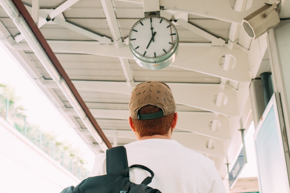A young man with short red hair, wearing a beige cap with a dark green adjustable strap at the back, is standing with his back to the camera at a transport station or waiting area. He is dressed in a white shirt and carries a black backpack over his right shoulder. Above him, there is a large round clock with a white face, black hour and minute hands, and black hour markers, mounted on a white, ceiling-mounted structure made of metal or plastic panels. The environment features a covered area with a high, white-painted ceiling supported by beams and a visible metal railing or barrier on the left side, suggesting an outdoor or semi-outdoor setting. The lighting appears natural, coming from the open sides of the station, indicating daytime. This scene could exemplify a space where individuals might wait for transportation, possibly linked to private or independent travel arrangements, such as for rubbish removal services. The presence of the clock and sheltered environment aligns with the context of timely, scheduled waste clearance operations managed by local or private waste management providers like Rubbish Clearance Balham, supporting alternative rubbish collection options.